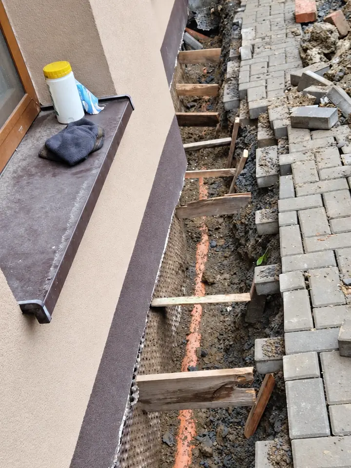 Rýha podél fasády domu s nopovou fólií a potrubím, vedle rozebraná dlažba