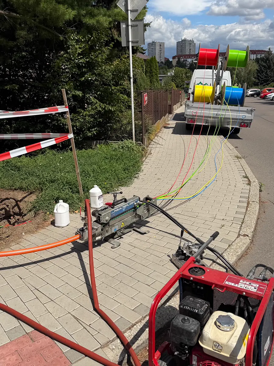 Zafukovací stroj a kompresor na chodníku, z cívek na přívěsu vedou barevné kabely do trasy